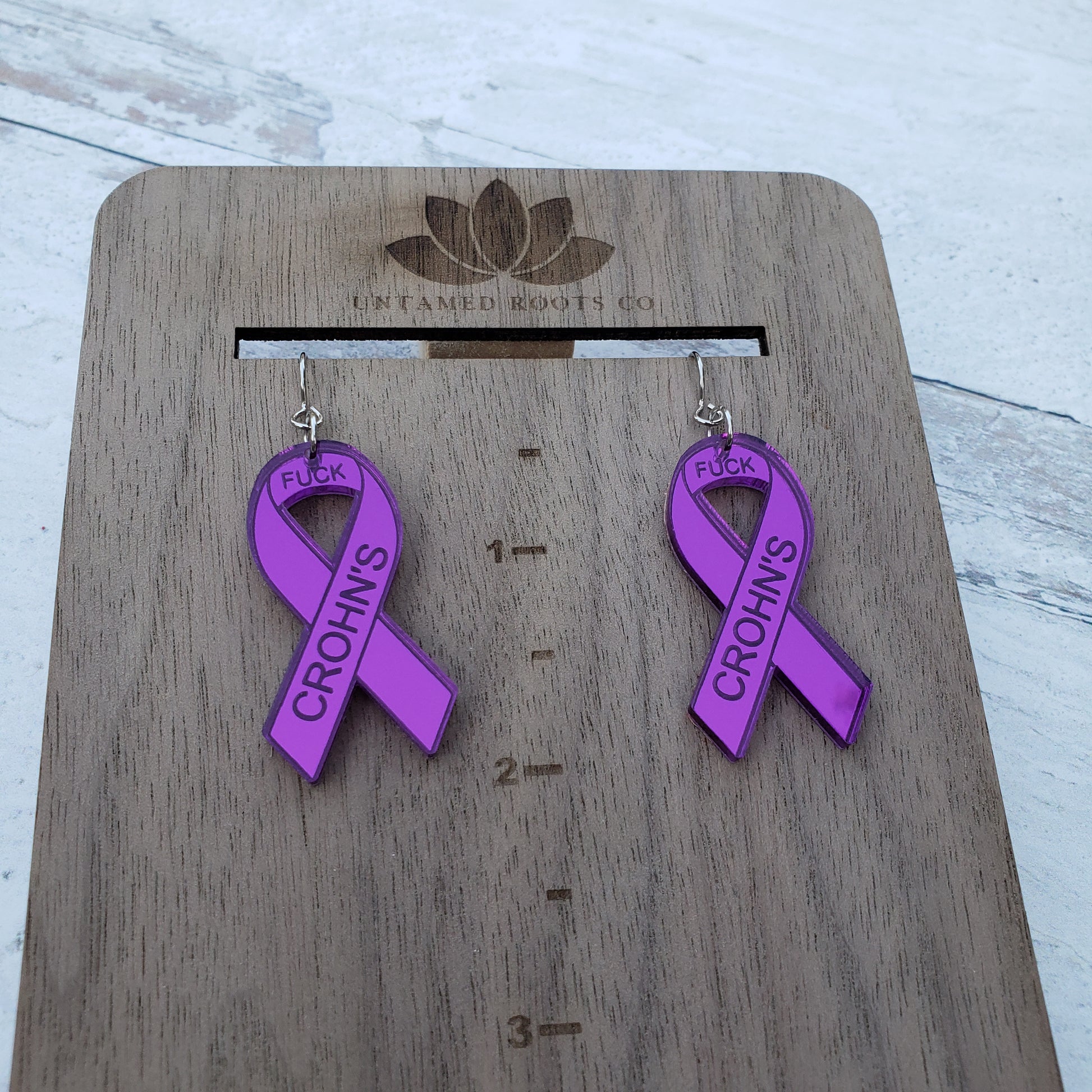 Fuck Crohn's Engraved purple mirror acrylic awareness ribbons on stainless steel earring wires. On hanging display for size.